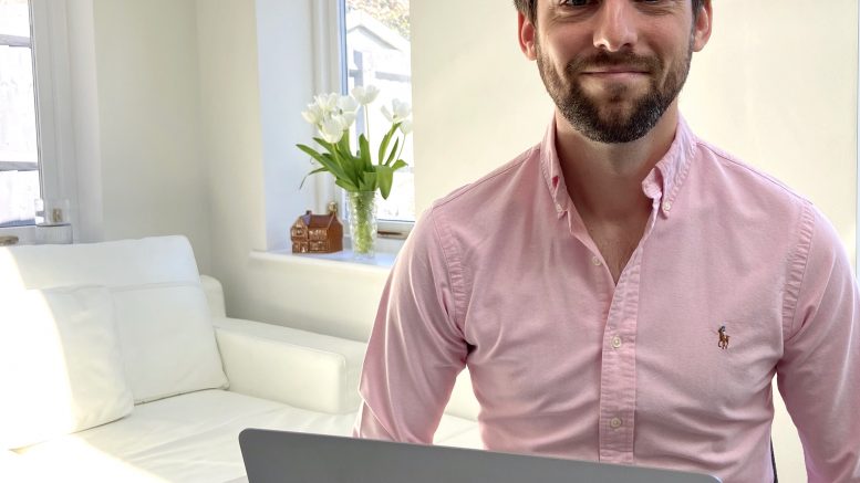 Jon Brown, in a pink shirt sat at a table with his silver laptop, a white sofa nearby.