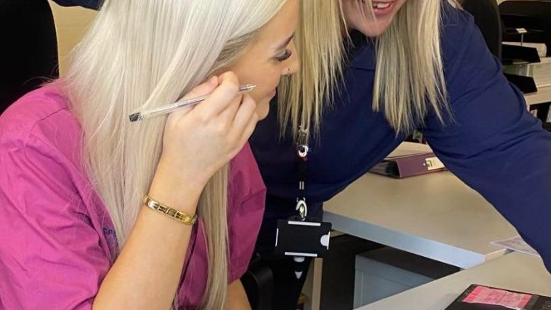 Two people, blonde ladies, one in a pink top and the other blue, sitting working at a desk.
