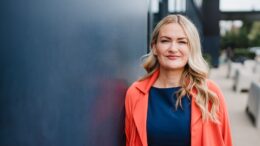 Picture of a smiling woman with blonde hair, blue top, and red jacket leaning against a blue wall.
